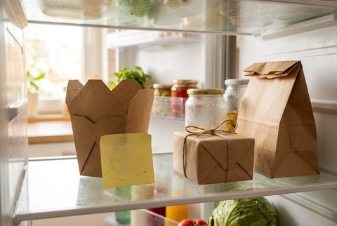 Open fridge with a takeout carton, a paper lunch bag, jars, a sticky note and a wrapped gift on the shelf