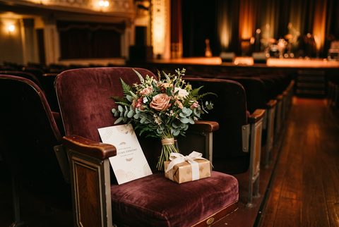Empty velvet theater seat holding a bouquet of flowers, a card and a small wrapped gift, with a stage in the background