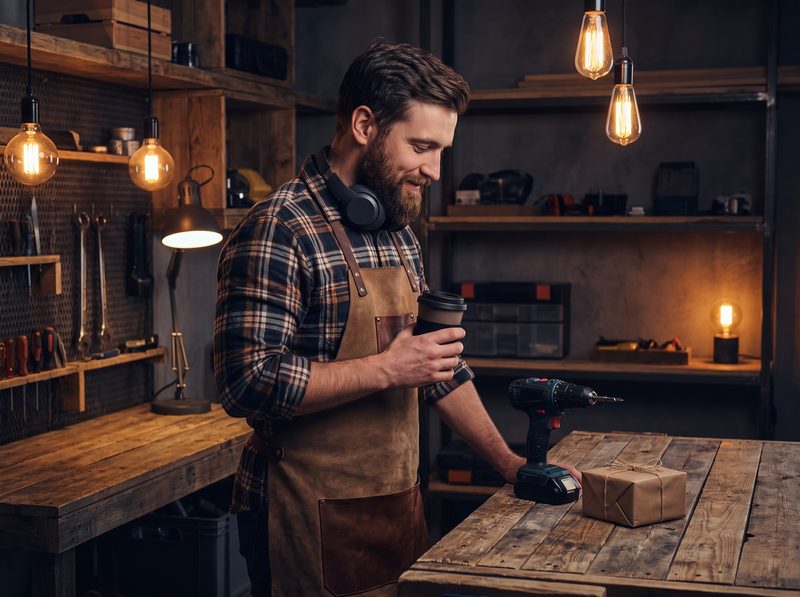 Man in a flannel shirt in a warm workshop holding a travel mug, headphones around his neck
