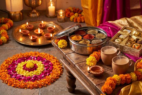 Diwali scene with glowing clay diyas on a brass tray, a flower rangoli, a steel masala dabba of spices and cups of chai