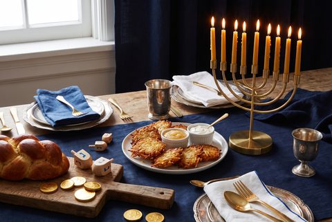 Hanukkah table with a brass menorah of lit candles, golden latkes with applesauce and sour cream, wooden dreidels, gelt and a braided challah