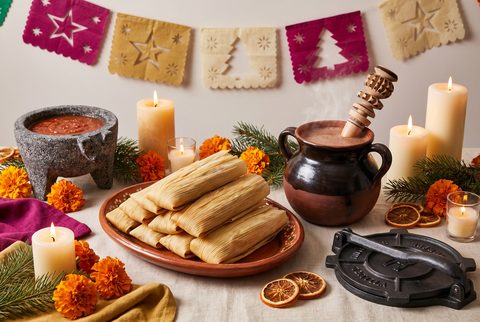 Nochebuena table with tamales on a clay platter, a pot of hot chocolate with a wooden molinillo, a molcajete of salsa and papel picado