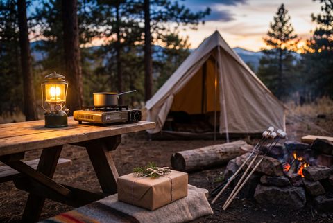 Campsite at dusk with a canvas tent, a glowing lantern and camp stove on a picnic table, a fire with roasting sticks and a wrapped gift