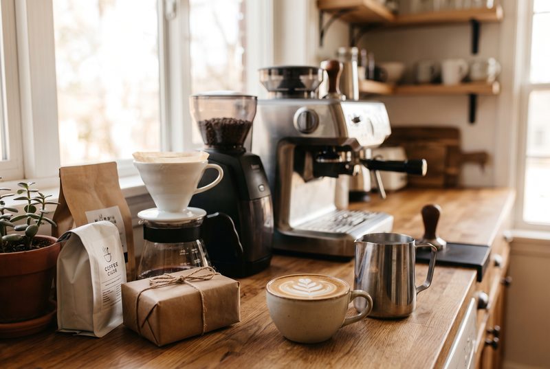 Kitchen coffee station with an espresso machine, a grinder, a pour-over dripper, a milk pitcher, a latte and a wrapped gift