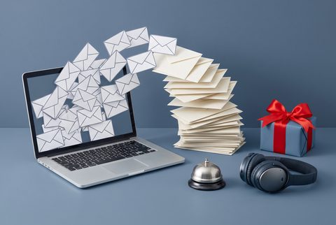 Laptop spewing a stream of envelopes beside a desk bell, headphones and a wrapped gift
