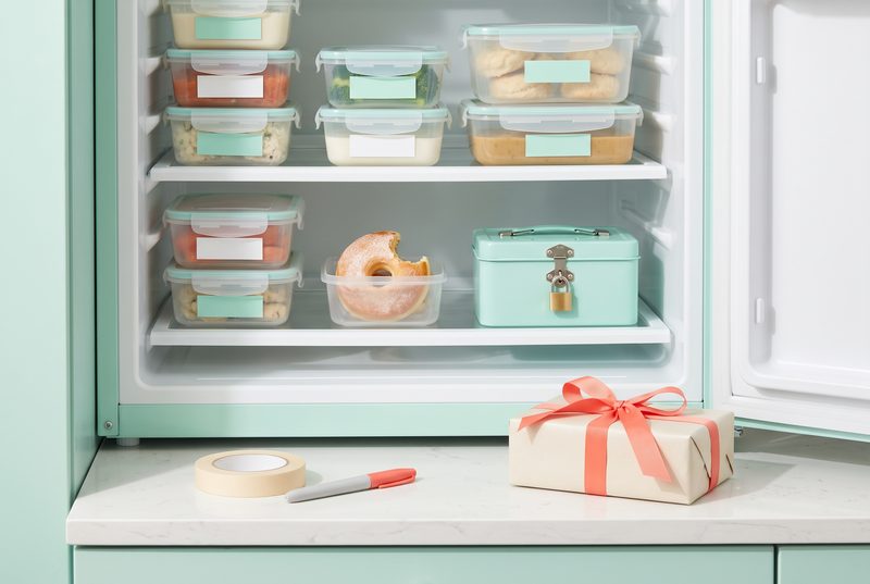 Open refrigerator full of labeled food containers, with a roll of tape and a wrapped gift