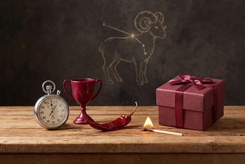 Wooden table with a stopwatch, a red trophy cup, a red chili pepper and a red wrapped gift beneath a ram constellation