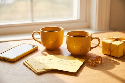 Two yellow mugs on a sunny table with a phone, handwritten note cards, dice and a yellow wrapped gift