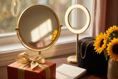 Gold vanity mirror and small ring light by a window with sunflowers, a notebook and an orange wrapped gift with gold ribbon