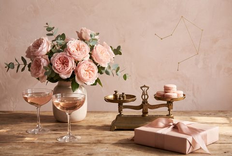 Pink roses in a vase on a wooden table with rosy coupes, a brass balance scale holding macarons and a pink wrapped gift