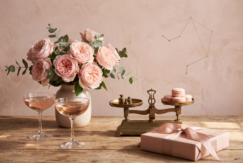 Pink roses in a vase on a wooden table with rosy coupes, a brass balance scale holding macarons and a pink wrapped gift
