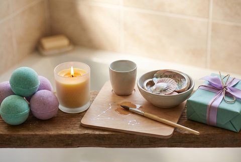Bathtub ledge with pastel bath bombs, a lit candle, a cup, a small bowl, a paintbrush on a board and a teal wrapped gift