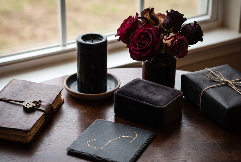Dark wooden table with a journal with a lock, a black candle, dark roses and a black wrapped gift