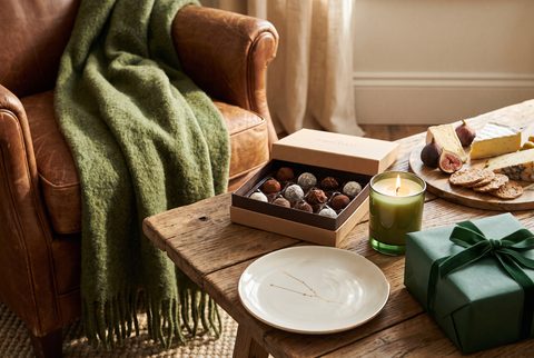 Leather armchair with a green knit throw beside chocolates, a candle, a cheese board and a green wrapped gift
