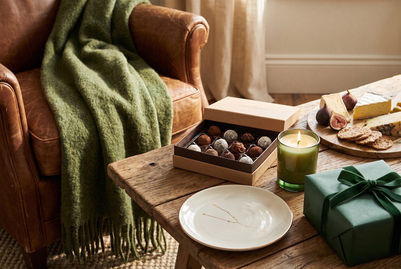 Leather armchair with a green knit throw beside chocolates, a candle, a cheese board and a green wrapped gift